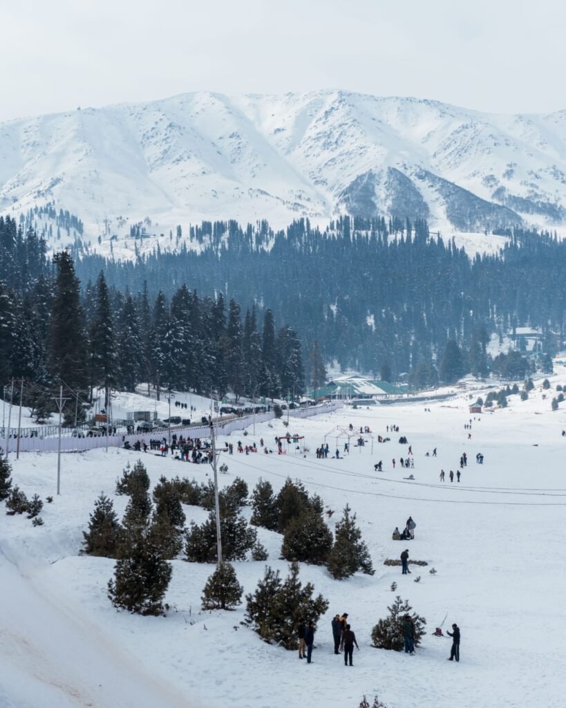 Scenic winter view of Gulmarg, India, with snowy mountains, pine trees, and people enjoying the outdoors.